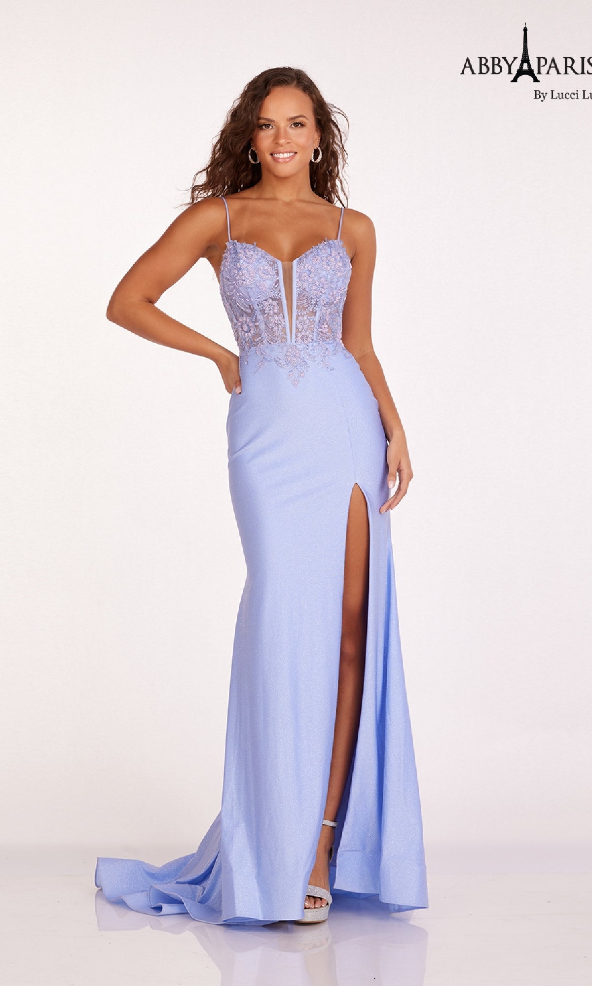 A woman wears the Sheer Corset Glitter Prom Dress Abby Paris 90207—light blue, embroidered, with thin straps, lace bodice, high slit, and floor-length hem—standing against a plain white background.